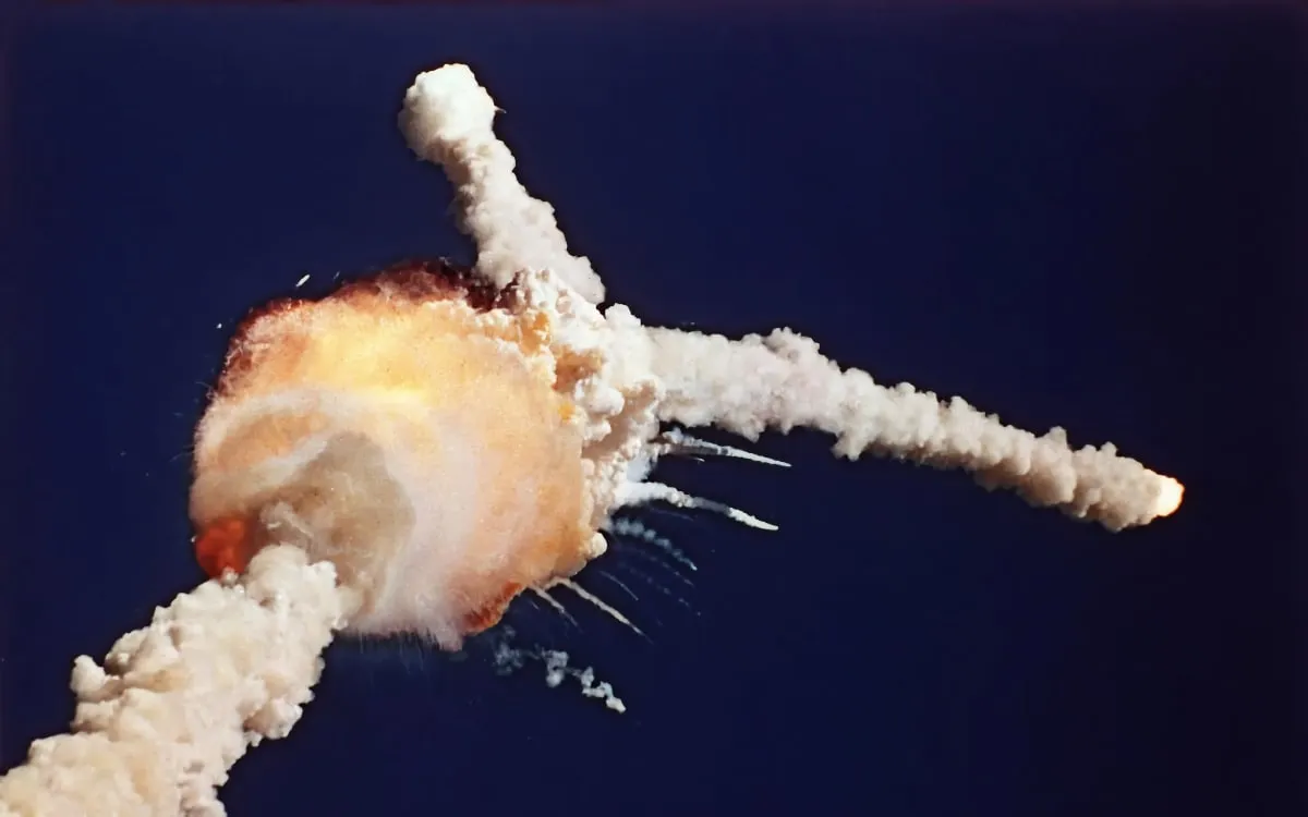 An orange fireball and billowing white cloud against a dark blue sky, with thick white trails branching upward and to the right and smaller streaks of debris spreading outward.