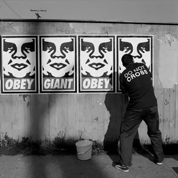 A black-and-white photo of a street artist pasting posters on a concrete wall. They say “Obey” and “Giant”