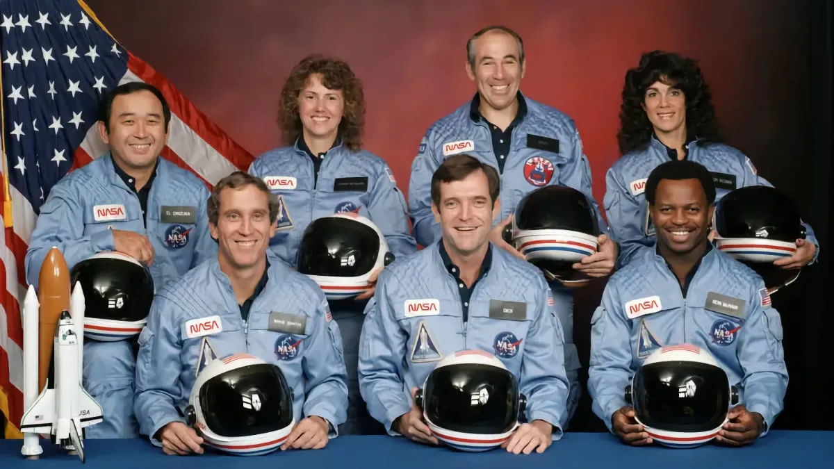 Seven astronauts posed next to a U.S. flag and a model of the space shuttle